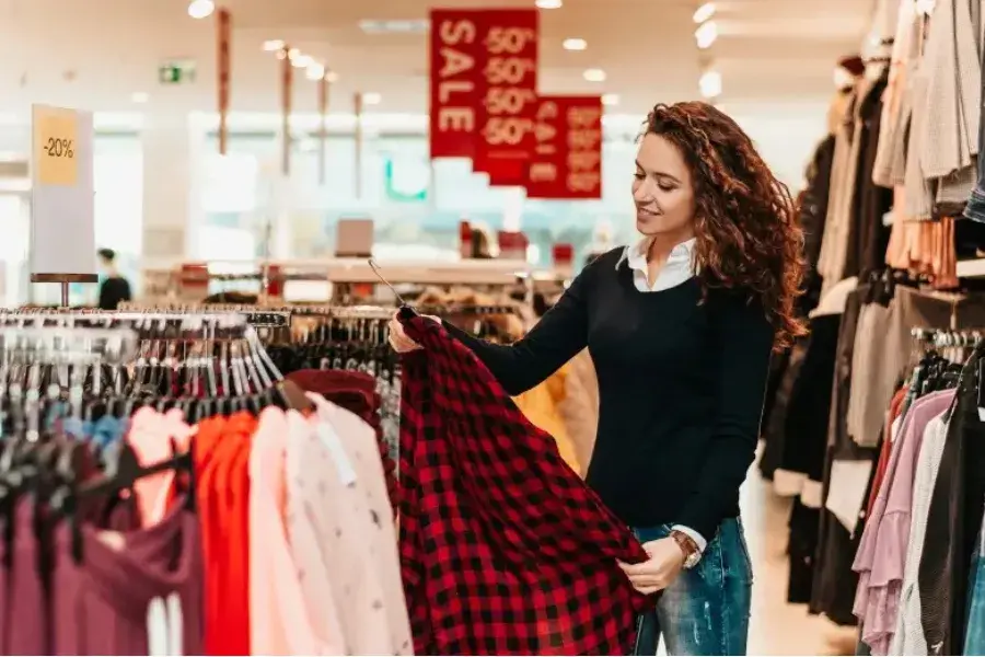 Woman shopping for clothes in a retail store holding a red plaid shirt with sale signs in background