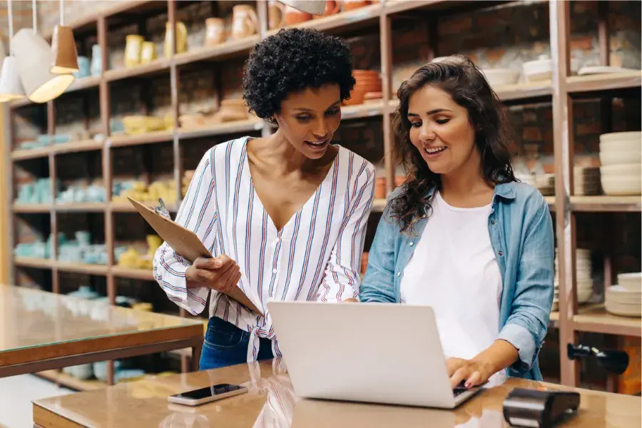 Two retail professionals reviewing sales data on a laptop, illustrating merchandise assortment planning for customer demand, seasonal trends, and inventory management.