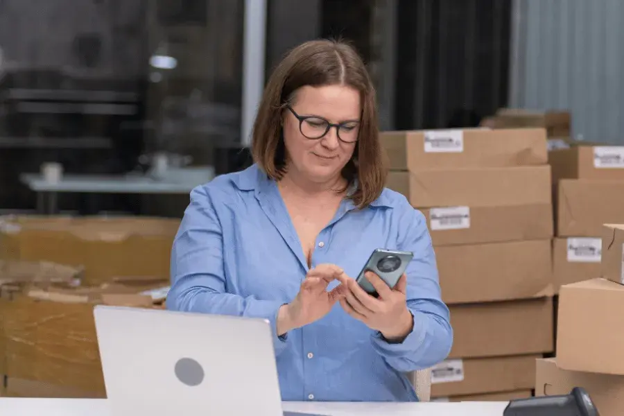 A woman retail business professional looking at her phone and laptop, with cardboard boxes stacked behind her — symbolizing sales and inventory management.