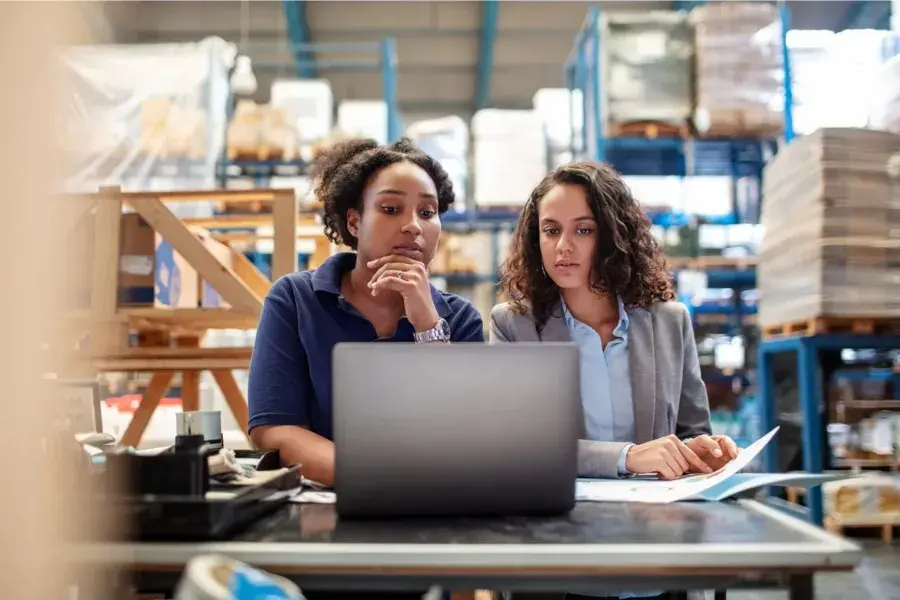 Two retail professionals reviewing inventory data on a laptop in a warehouse, illustrating real-time merchandise financial planning, scenario planning, and inventory optimization.