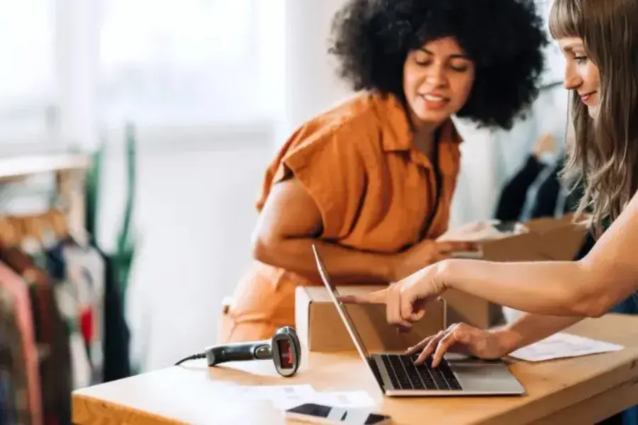 Two women reviewing data on a laptop while packing boxes, illustrating AI-powered assortment planning, demand forecasting, SKU management, and data-driven retail decision making.