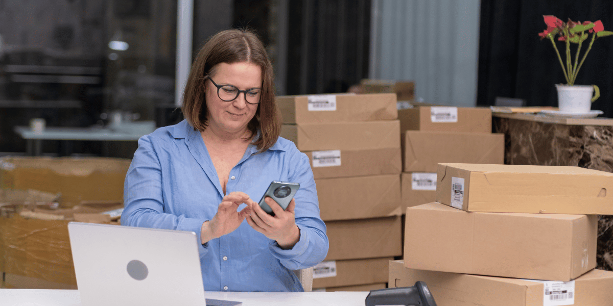 A woman retail business professional looking at her phone and laptop, with cardboard boxes stacked behind her — symbolizing sales and inventory management.