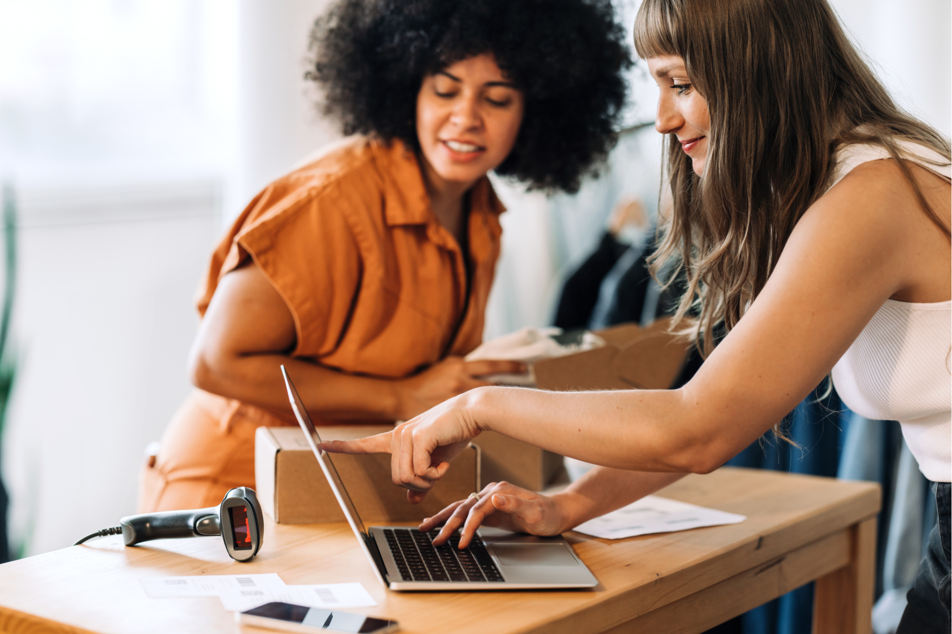 Two women reviewing data on a laptop while packing boxes, illustrating AI-powered assortment planning, demand forecasting, SKU management, and data-driven retail decision making.