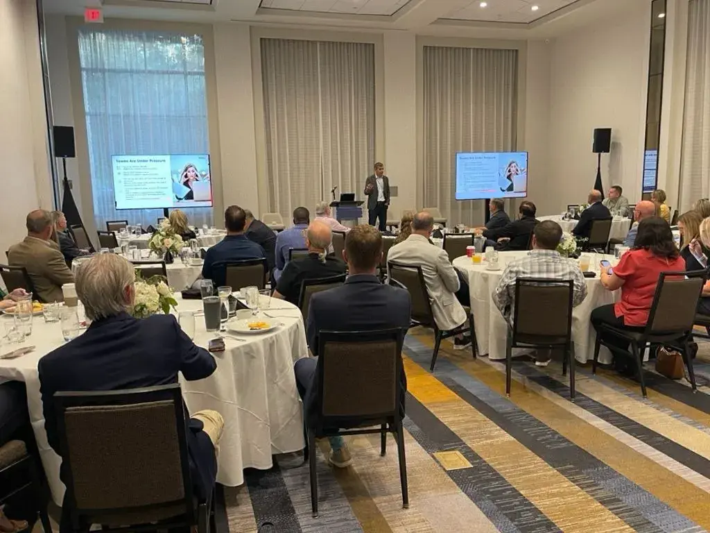 Speaker presenting the Hub Model to retail partners during a business summit, with attendees seated around tables in a conference room.